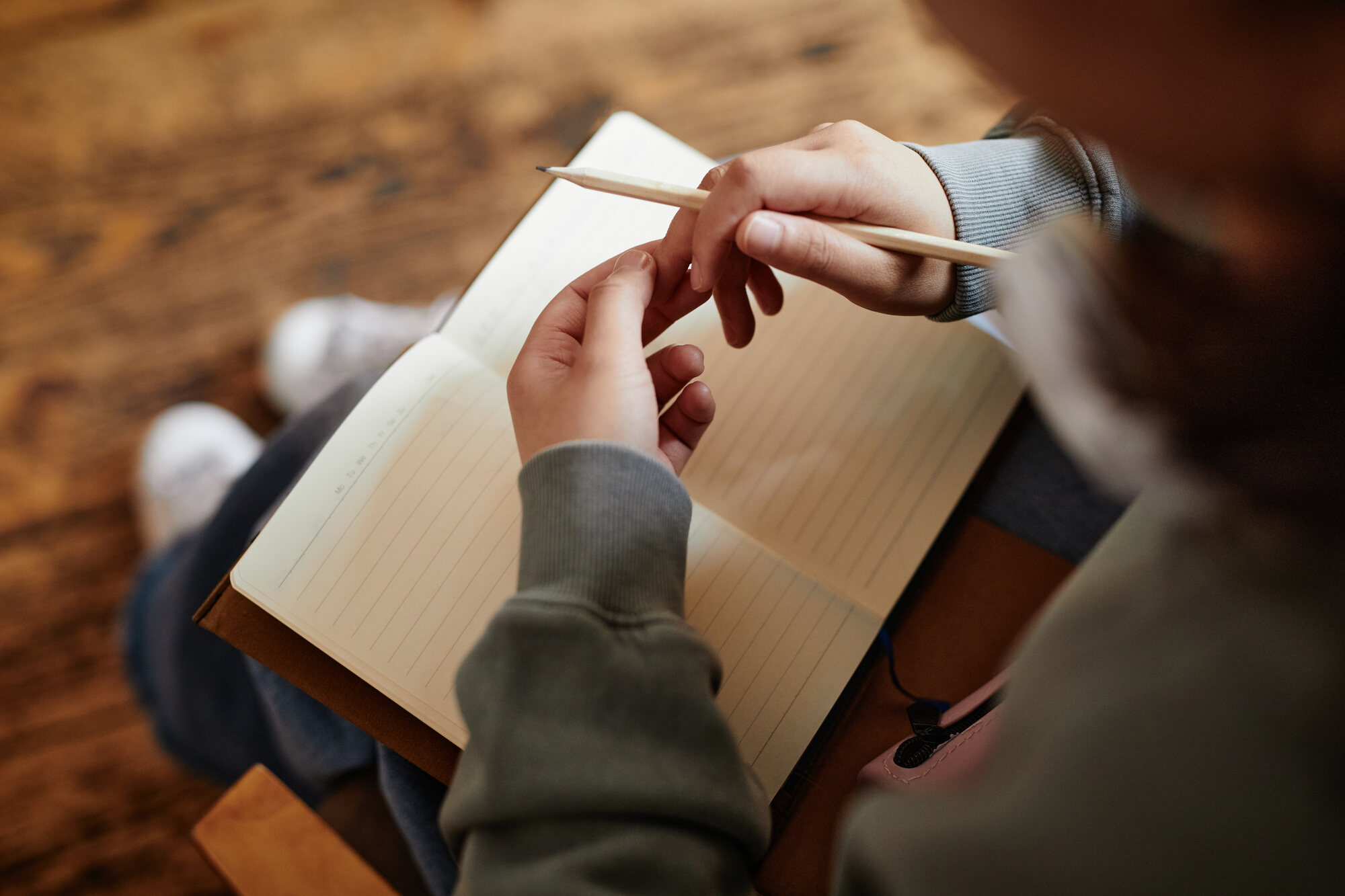 A person writing in an open notebook while seated on a wooden floor